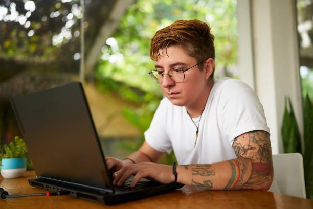 Focused individual with tattoos tech for remote job. Transgender freelancer works on laptop in tropical coworking space. Open, inclusive work plants. Creativity, productivity LGBTQ-friendly setting.